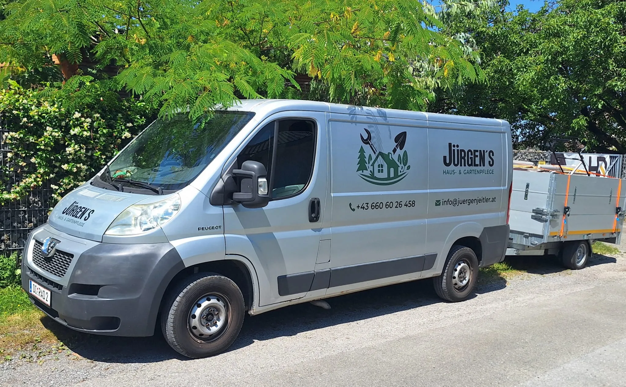 Firmenwagen mit Anhänger von Jürgen's Haus- & Gartenpflege unter einem Baum in Dornbirn